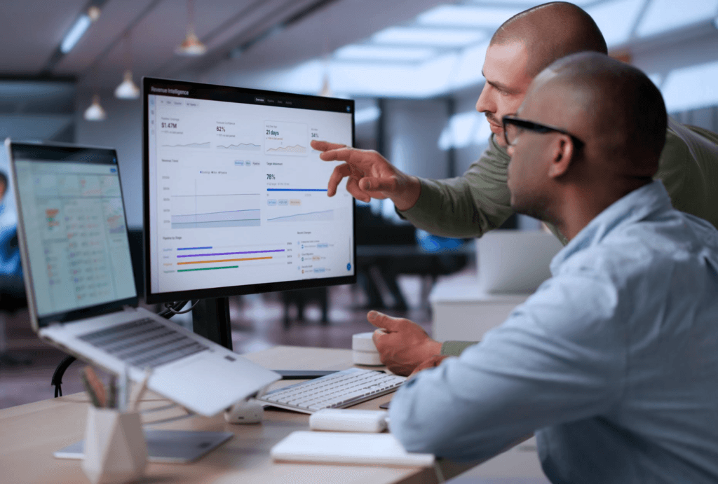 Two professional colleagues reviewing a business intelligence and revenue analytics dashboard on a desktop monitor in a modern office